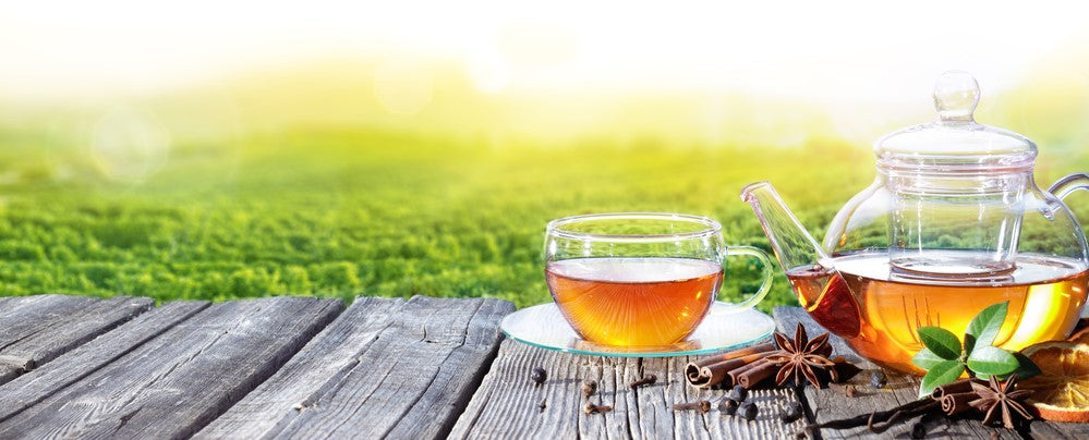 Tea set with a glass cup, teapot and some herbs lying besides star anise, cinnamon, juniper, cloves orange vanilla tea leaves. On a wooden table with a blurred tea plantation in the background